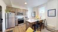 Located on Colerain Avenue in Cincinnati, Ohio, this thoughtfully designed one-bedroom, one-bathroom unit makes excellent use of its compact footprint across a single floor. The open-concept living and kitchen area serves as the heart of the home, featuring warm wood-toned cabinetry, stainless steel appliances including a refrigerator, range, and microwave, and a white quartz peninsula with bar seating — ideal for casual dining or working from home. Light wood-look flooring flows throughout the main living space, complemented by crisp white walls that keep the interior feeling bright and airy. Natural light filters in through windows fitted with blinds. The bedroom offers a cozy retreat with carpeted floors, dual windows dressed in blue velvet curtains, and a built-in closet for ample storage. A stacked washer and dryer unit is conveniently located within the bedroom area. The bathroom is a standout feature, showcasing bold black-and-white patterned encaustic floor tile, marble-style shower surround with a glass sliding door, gold hardware accents, and a dark wood vanity — a refined combination of classic and contemporary design. This well-appointed unit delivers modern comfort and practical living in one cohesive space.