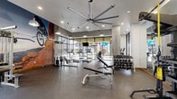 Located at 350 E Congress St in downtown Tucson, AZ, this contemporary fitness center delivers a well-rounded workout experience within a thoughtfully designed, single-floor space. The room is anchored by a striking oversized industrial ceiling fan that complements the modern aesthetic, while recessed lighting and pendant fixtures ensure consistent, bright illumination throughout.

Floor-to-ceiling glass windows line one side of the facility, flooding the interior with natural light and offering views of the surrounding streetscape. The durable rubber flooring provides a safe, high-performance surface across all workout zones. A bold, large-scale mural of a mountain biker navigating red rock terrain serves as a dynamic focal point, energizing the atmosphere and reinforcing an active lifestyle theme.

The cardio section features a row of treadmills and elliptical trainers, each equipped with individual monitors for entertainment during workouts. The strength training area is comprehensively outfitted with a cable machine system, a full rack of dumbbells, adjustable benches, kettlebells, medicine balls, and resistance training accessories. Full-length mirrors extend across key walls, enhancing the sense of openness and supporting proper form. Exposed concrete columns contribute an industrial character, while glass-panel partitions define zones without sacrificing the airy, open feel of the space.