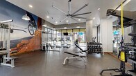 Located at 350 E Congress St in downtown Tucson, AZ, this contemporary fitness center delivers a well-rounded workout experience within a thoughtfully designed, single-floor space. The room is anchored by a striking oversized industrial ceiling fan that complements the modern aesthetic, while recessed lighting and pendant fixtures ensure consistent, bright illumination throughout.

Floor-to-ceiling glass windows line one side of the facility, flooding the interior with natural light and offering views of the surrounding streetscape. The durable rubber flooring provides a safe, high-performance surface across all workout zones. A bold, large-scale mural of a mountain biker navigating red rock terrain serves as a dynamic focal point, energizing the atmosphere and reinforcing an active lifestyle theme.

The cardio section features a row of treadmills and elliptical trainers, each equipped with individual monitors for entertainment during workouts. The strength training area is comprehensively outfitted with a cable machine system, a full rack of dumbbells, adjustable benches, kettlebells, medicine balls, and resistance training accessories. Full-length mirrors extend across key walls, enhancing the sense of openness and supporting proper form. Exposed concrete columns contribute an industrial character, while glass-panel partitions define zones without sacrificing the airy, open feel of the space.