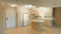 This single-floor space centers around a bright, functional kitchen that serves as its defining feature. The kitchen is outfitted with an abundance of light maple cabinetry, providing generous storage across both upper and lower cabinets. A central island with a grey laminate countertop anchors the space, offering additional prep area and casual seating potential. The kitchen is fully equipped with white appliances, including a full-size refrigerator, electric range, built-in microwave, and dishwasher, all seamlessly integrated into the cabinetry layout. Track lighting mounted on the ceiling illuminates the workspace efficiently, highlighting the clean lines of the cabinetry and countertops. The sink is positioned along the perimeter counter, with a view toward the open kitchen area. Light hardwood-style flooring runs throughout, lending warmth and continuity to the space. The overall aesthetic is clean and understated, with neutral tones throughout the cabinetry, countertops, and walls creating a cohesive and inviting environment. The open layout between the island and the surrounding counters allows for easy movement and a practical workflow, making this a highly functional and well-designed kitchen space.
