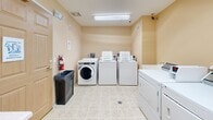 This shared laundry facility at 770 Savanna Ave in Saint Cloud, Minnesota, is a compact, well-organized space designed to serve the practical needs of its users. The room features warm beige walls and durable light-toned tile flooring that is easy to maintain and clean. Overhead fluorescent lighting ensures the space is well-lit throughout.

The laundry room is equipped with multiple commercial-grade top-loading and front-loading washing machines, as well as a matching set of dryers, providing ample capacity for residents. A wall-mounted coin-operated vending machine offers convenient access to laundry detergent and other supplies. A built-in folding counter with an angled corner design runs along one wall, offering a generous surface for sorting and folding laundry. A utility sink is also available for hand-washing or pre-treating garments.

Practical amenities include a wall-mounted flat-screen television, providing entertainment while waiting for laundry to complete. A fire extinguisher and trash receptacle are positioned near the entry door, reflecting attention to safety and cleanliness. Ventilation is provided through ceiling and wall-mounted vents, maintaining air circulation throughout the space. A window allows for a modest amount of natural light. The entry features a solid wood-panel door, ensuring privacy and security for users.