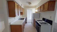 This single-floor unit offers a thoughtfully organized layout with distinct living, sleeping, and functional zones. The galley-style kitchen is efficiently designed with warm honey-toned wood cabinetry lining both walls, providing ample upper and lower storage. White laminate countertops offer generous prep space, complemented by a stainless steel sink, a gas range, a dishwasher, and a full-size refrigerator. Light vinyl tile flooring and a sliding glass door at the far end of the kitchen allow natural light to filter in. The open living area is carpeted in neutral tones and features a wide vertical blind-covered sliding door, lending the space an airy, bright feel. The bedroom spaces are also carpeted and neutral in palette, with one bedroom showcasing built-in sliding closet doors and direct access to the entry. Both bathrooms are finished with white mosaic tile flooring, a clean white vanity with dark espresso cabinetry, a mirror, and updated fixtures. One bathroom includes a full bathtub with shower. Throughout, the space maintains a clean, neutral aesthetic with light walls and consistent finishes, offering a move-in-ready environment suited to a variety of lifestyles.