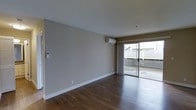 This single-floor space features a clean, minimalist design with a focus on functionality and natural light. The main living area is finished with rich, dark hardwood flooring that runs throughout, providing a warm contrast to the neutral-toned walls painted in a soft greige. White baseboards frame the perimeter, adding a crisp, polished detail to the room.

A wall-mounted mini-split air conditioning unit is positioned near the ceiling, offering efficient climate control without occupying floor space. A wall-mounted intercom or control panel is also visible, adding a practical convenience feature to the space.

The most prominent feature of the living area is the large sliding glass door, framed by floor-length grey curtains. This door opens to an outdoor patio or terrace, enclosed by a white lattice fence, providing a semi-private outdoor extension of the living space. Natural light filters generously through the glass, brightening the interior throughout the day.

The overall ambiance is open and airy, with a neutral palette that creates a versatile backdrop suitable for a variety of interior styles. The space presents a straightforward, well-maintained layout with practical features throughout.