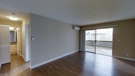 This single-floor space features a clean, minimalist design with a focus on functionality and natural light. The main living area is finished with rich, dark hardwood flooring that runs throughout, providing a warm contrast to the neutral-toned walls painted in a soft greige. White baseboards frame the perimeter, adding a crisp, polished detail to the room.

A wall-mounted mini-split air conditioning unit is positioned near the ceiling, offering efficient climate control without occupying floor space. A wall-mounted intercom or control panel is also visible, adding a practical convenience feature to the space.

The most prominent feature of the living area is the large sliding glass door, framed by floor-length grey curtains. This door opens to an outdoor patio or terrace, enclosed by a white lattice fence, providing a semi-private outdoor extension of the living space. Natural light filters generously through the glass, brightening the interior throughout the day.

The overall ambiance is open and airy, with a neutral palette that creates a versatile backdrop suitable for a variety of interior styles. The space presents a straightforward, well-maintained layout with practical features throughout.