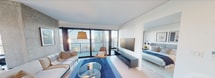 First Light 2409 is a modern high-rise residence offering 780 square feet of thoughtfully designed living space across 3 bedrooms, 2 bathrooms, and 1 closet on a single floor. The open-plan living and dining area is defined by expansive floor-to-ceiling sliding glass doors that flood the interior with natural light and frame stunning panoramic views of the city skyline and waterfront. The dining area features pendant lighting with brass accents, a full-length dining table, and direct access to a wraparound glass-railed balcony offering sweeping elevated views of the surrounding urban landscape. The living room is anchored by large-format windows, light hardwood flooring, and clean white walls that create a bright, contemporary atmosphere. The primary bedroom, at 100 square feet, includes direct balcony access through floor-to-ceiling glazing, providing exceptional city views. The bathrooms are finished with floor-to-ceiling marble-style stone tiling, a soaking tub, rain shower head, and matte black fixtures throughout, lending a spa-like quality. The unit's minimalist design is consistent throughout, with white walls, recessed lighting, and light flooring creating a cohesive, sophisticated aesthetic.