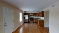 This single-story space showcases an open-concept layout where the kitchen serves as a central focal point. The kitchen is outfitted with warm-toned wooden cabinetry that extends to the upper walls, providing generous storage throughout. Granite countertops in earthy tones complement the cabinetry, while stainless steel appliances—including a French door refrigerator with an ice and water dispenser, a range with an electric cooktop, and an over-the-range microwave—add a modern, functional touch. A double sink is positioned along the far counter, benefiting from natural light streaming through a nearby window fitted with horizontal blinds. Hardwood flooring in a rich, medium-toned finish runs seamlessly throughout the visible areas, adding warmth and cohesion to the space. The open floor plan creates a sense of spaciousness, with the kitchen flowing naturally into the adjoining living area. Neutral-toned walls and white ceilings keep the interior bright and versatile, offering a clean backdrop that accommodates a variety of décor styles. The overall ambiance is practical yet inviting.