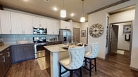This thoughtfully designed single-floor residence combines modern finishes with a warm, sophisticated aesthetic throughout. The chef-inspired kitchen is a standout feature, showcasing a two-tone cabinetry scheme of crisp white uppers and rich dark wood lowers, a subway tile backsplash, stainless steel appliances, and a granite-topped island with seating for casual dining. Pendant lighting illuminates the space, adding an inviting glow. The living area offers a comfortable and stylish retreat, with crown molding, a ceiling fan, hardwood-style flooring, and a glass door that opens to a private balcony, filling the room with natural light. The primary bedroom is generously proportioned, featuring plush carpet, a ceiling fan, and ample room for a seating area. A secondary bedroom provides a cozy and intimate atmosphere. The primary bathroom impresses with a dual-sink vanity, granite countertops, dark wood cabinetry, a soaking tub, and a separate shower. A second bathroom echoes the same refined design with its own granite vanity and framed mirror. Consistent hardwood-style flooring, crown molding, and a neutral yet sophisticated color palette tie the entire residence together seamlessly.