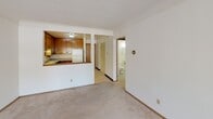 Located at 2297 Standish St in the Hamline-Midway neighborhood of Saint Paul, Minnesota, this single-floor unit offers a practical and well-organized layout. The living area is generously sized, featuring light-toned carpeting, white walls, and a large window with vertical blinds that allows natural light to fill the room. A wall-mounted air conditioning unit and baseboard heating provide year-round climate control. The kitchen is accessed via a half-wall pass-through from the living area, creating an open connection between the two spaces. It features warm-toned wood cabinetry, speckled laminate countertops, a double-basin stainless steel sink, and white appliances including a range and refrigerator. A dedicated bedroom offers carpeted flooring, a window with a baseboard heater beneath it, and a built-in closet with louvered doors providing ample storage with a hanging rod and shelf. The full bathroom is finished with white square wall tile surrounding the tub-shower combination, a mosaic tile floor, a pedestal sink, and a large mirror. A hallway connects the main living spaces to the bedroom and bathroom, maintaining a clear and functional flow throughout the unit.
