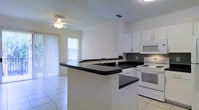 This three-floor space features an open-concept layout that seamlessly connects the kitchen and living area, creating a versatile and functional environment. The kitchen is equipped with white cabinetry, white appliances including a range and built-in microwave, and a wraparound peninsula countertop finished in dark laminate. The peninsula integrates a sink and provides additional prep and seating space, making it a central hub of the layout.

Light tile flooring extends throughout the main living area, reflecting natural light and contributing to the bright, airy atmosphere. A ceiling fan is centrally positioned overhead, enhancing airflow and comfort. The living area benefits from large sliding glass doors fitted with vertical blinds, which open to a balcony overlooking lush greenery, bringing the outdoors in and providing pleasant views. An additional window further supplements the natural light in the space.

The clean white walls and neutral palette throughout give the space a fresh, open feel, while the textured ceiling adds a subtle architectural detail. The overall design prioritizes functionality and light, making it a well-appointed, practical living environment spread across three floors.