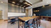This thoughtfully designed single-floor space functions as a communal lounge and collaborative hub, combining multiple distinct seating areas within an open, well-appointed layout. The entryway features a striking herringbone-patterned wood tile floor transitioning to large-format light tile, setting a refined tone upon arrival. A clearly marked meeting room with a glass-paneled black door offers a semi-private workspace alongside the main lounge.

The central gathering area is anchored by a substantial natural wood communal table with counter-height seating, complemented by upholstered blue chairs with walnut-toned frames. Overhead, a decorative geometric lattice screen in dark metal adds architectural depth, paired with a sleek pendant light fixture. A built-in banquette with soft gray upholstery runs along one wall, providing additional casual seating alongside smaller bistro tables.

A secondary lounge zone features a tufted leather curved banquette, black oval tables, and bold artwork, beneath tall ceilings with drum pendant lighting. Along the windowed perimeter, a long counter-height ledge with individual seating faces large windows, flooding the space with natural light and offering views of the surrounding exterior. A dark navy textured accent wall with sculptural sconces adds a striking design focal point throughout.