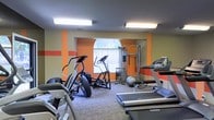 This fitness center is designed to support a well-rounded workout experience within a single-floor layout. The room features bold orange accent stripes set against neutral beige walls, giving the space an energetic and motivating atmosphere. Overhead fluorescent lighting ensures the area is well-lit throughout, complemented by a arched window along the back wall that allows natural light to filter in, offering a pleasant outdoor view of surrounding trees.

The cardio section is well-appointed, housing multiple treadmills, elliptical trainers, and a stair climber-style machine, each equipped with individual display consoles for tracking workout metrics. A multi-functional cable weight machine is positioned toward the back of the room, providing options for a variety of strength-training exercises. A stability ball and additional free weight equipment round out the strength training offerings.

The flooring consists of low-pile carpet, providing cushioning underfoot while maintaining a clean, functional aesthetic. The layout is thoughtfully arranged to allow adequate space between machines, ensuring comfortable movement throughout the facility.