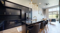 This contemporary single-floor residence showcases a thoughtfully curated design that balances bold sophistication with comfortable living. The standout kitchen is a true centerpiece, outfitted with floor-to-ceiling matte black cabinetry accented by brushed gold hardware, dark granite countertops, and a large island with an undermount sink and seating for multiple bar-height chairs upholstered in charcoal velvet. Cascading crystal pendant lights overhead add a glamorous finishing touch, while stainless steel appliances — including a built-in microwave, range, and French door refrigerator — provide full functionality. Light oak hardwood flooring runs throughout the main living areas, offering a warm contrast to the kitchen's dramatic palette. The living room impresses with soaring ceilings and a floor-to-ceiling textured fireplace wall that serves as a dramatic focal point, complemented by generous natural light. The bedroom continues the airy feel with towering ceilings, large windows framing lush green outdoor views, soft carpeting, and warm wood-toned interior doors. A walk-in closet adds practical storage. The bathroom features light wood cabinetry, a marble-topped vanity, and clean hardwood-style flooring, completing this well-appointed and design-forward space.