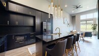 This contemporary single-floor residence showcases a thoughtfully curated design that balances bold sophistication with comfortable living. The standout kitchen is a true centerpiece, outfitted with floor-to-ceiling matte black cabinetry accented by brushed gold hardware, dark granite countertops, and a large island with an undermount sink and seating for multiple bar-height chairs upholstered in charcoal velvet. Cascading crystal pendant lights overhead add a glamorous finishing touch, while stainless steel appliances — including a built-in microwave, range, and French door refrigerator — provide full functionality. Light oak hardwood flooring runs throughout the main living areas, offering a warm contrast to the kitchen's dramatic palette. The living room impresses with soaring ceilings and a floor-to-ceiling textured fireplace wall that serves as a dramatic focal point, complemented by generous natural light. The bedroom continues the airy feel with towering ceilings, large windows framing lush green outdoor views, soft carpeting, and warm wood-toned interior doors. A walk-in closet adds practical storage. The bathroom features light wood cabinetry, a marble-topped vanity, and clean hardwood-style flooring, completing this well-appointed and design-forward space.