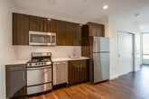 This contemporary single-floor residence showcases a thoughtfully designed open layout with rich hardwood flooring running throughout. The kitchen is a standout feature, outfitted with dark espresso-toned cabinetry accented by a bold wood-grain finish, crisp white countertops, and a classic subway tile backsplash. A full suite of stainless steel appliances—including a refrigerator, range, microwave, and dishwasher—provides a polished, modern aesthetic. Recessed lighting illuminates the kitchen and living areas, enhancing the clean, contemporary feel. The living area is generously proportioned, offering an open canvas bathed in natural light streaming through floor-to-ceiling windows. These expansive windows frame striking panoramic views of the surrounding urban landscape, including what appears to be a notable civic structure in the distance. The bathroom continues the modern theme with a glass-enclosed walk-in shower clad in white subway tiles, chrome fixtures, and a built-in illuminated mirror, creating a spa-like atmosphere. A sliding door near the entry adds a space-efficient architectural touch. Throughout the residence, the consistent use of neutral tones, quality finishes, and abundant natural light create a bright, inviting, and cohesive living environment.