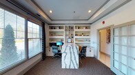 This single-floor office space is thoughtfully designed to balance functionality with visual appeal. The room's most striking feature is a central table with a bold marble-style surface, showcasing dramatic grey veining against a white background — an eye-catching focal point that doubles as a collaborative workspace. Flanking the room along the back wall is a built-in white shelving and desk unit, providing ample storage and dedicated workstations equipped with desktop computers and task chairs. The shelving displays a curated selection of decorative objects, books, and plants, adding warmth and character to the professional setting. A tray ceiling with recessed lighting adds architectural depth and a refined aesthetic overhead. Natural light filters in through a large window dressed with patterned curtains and horizontal blinds, brightening the carpeted interior. The neutral color palette of white, grey, and beige creates a clean and organized atmosphere, while a doorway leads to an adjacent room, suggesting this space is part of a larger facility. Overall, the space conveys a polished, professional environment suited for office or business center use.