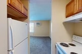 Located at 120 Polk St in Syracuse, NY, this one-bedroom, one-bathroom unit offers a practical and efficient single-floor layout. The bedroom is the largest space in the unit, featuring wall-to-wall carpeting, neutral beige walls, a window with outdoor views of surrounding greenery, a wall-mounted air conditioning unit, and baseboard heating — ensuring year-round comfort. A closet with a paneled door provides convenient storage within the bedroom. The open sightlines from the bedroom into the kitchen create a sense of connectivity throughout the unit. The kitchen is galley-style and includes warm-toned oak cabinetry, dark granite-look countertops, a stainless steel sink, a white electric coil range with an overhead vent hood, and a full-size refrigerator. The flooring in the kitchen transitions to hardwood-style planks, adding a touch of warmth. The bathroom is accessed via a short hallway and features wood-look plank flooring, a rich wood-toned vanity with a white countertop, a medicine cabinet mirror, and direct sightlines back to the kitchen. This unit is well-suited for those seeking a straightforward, functional living space in Syracuse's residential neighborhood.