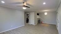 This single-story space offers an open, airy layout defined by clean lines and modern finishes throughout. The main living area features light gray walls complemented by crisp white crown molding and baseboards, creating a polished, cohesive aesthetic. Light-toned laminate flooring extends across the space, adding warmth while maintaining a contemporary feel.

A ceiling fan with a built-in light fixture anchors the living area, while recessed lighting provides additional illumination. A decorative pendant light near the dining area adds a stylish accent. The open floor plan connects the living space to a functional kitchen, visible through a pass-through opening equipped with a breakfast bar and bar stool seating—ideal for casual dining or entertaining.

The kitchen, partially visible from the main area, is outfitted with stainless steel appliances, including a range and microwave, set against dark cabinetry. A white paneled interior door provides access to an additional room, maintaining the clean, bright character of the space. Overall, the layout is practical and inviting, with thoughtful design details that balance comfort and modern style.