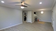 This single-story space offers an open, airy layout defined by clean lines and modern finishes throughout. The main living area features light gray walls complemented by crisp white crown molding and baseboards, creating a polished, cohesive aesthetic. Light-toned laminate flooring extends across the space, adding warmth while maintaining a contemporary feel.

A ceiling fan with a built-in light fixture anchors the living area, while recessed lighting provides additional illumination. A decorative pendant light near the dining area adds a stylish accent. The open floor plan connects the living space to a functional kitchen, visible through a pass-through opening equipped with a breakfast bar and bar stool seating—ideal for casual dining or entertaining.

The kitchen, partially visible from the main area, is outfitted with stainless steel appliances, including a range and microwave, set against dark cabinetry. A white paneled interior door provides access to an additional room, maintaining the clean, bright character of the space. Overall, the layout is practical and inviting, with thoughtful design details that balance comfort and modern style.