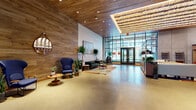 This expansive lobby makes a bold architectural statement through a thoughtful fusion of industrial and organic materials. Warm wood-planked ceilings span the length of the space, complemented by polished concrete floors that reflect abundant natural light. A dramatic skylight framed in steel and adorned with a sculptural hanging installation of layered wood elements serves as a striking focal point overhead.

Floor-to-ceiling steel-framed glass walls and double-door entries flood the interior with daylight and provide views to the surrounding streetscape. Exposed concrete columns and accent walls add an industrial edge, while live-edge wood benches and recessed floor lighting introduce refined, tactile details.

The lobby is organized into distinct seating areas furnished with bold, mid-century-inspired pieces — including deep emerald tufted sofas, sculptural high-back blue armchairs, and patterned area rugs — all accented by an abundance of lush indoor plants. Curated artwork adorns the walls, lending a gallery-like quality to the environment.

A reception desk and open shelving unit anchor one side of the space, providing functional counterbalance to the lounge areas. Throughout, the design achieves a warm yet contemporary ambiance, balancing raw materiality with carefully chosen furnishings to create a welcoming and visually dynamic arrival experience.
