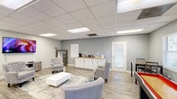 Located at 276 Upper Riverdale Rd in Jonesboro, Georgia, this single-floor commercial space offers a thoughtfully designed open-plan layout that combines comfort and functionality. The main room features light gray walls, wood-look luxury vinyl plank flooring, and a drop ceiling with recessed panel lighting that creates a bright, welcoming ambiance throughout.

The lounge area serves as the focal point of the space, furnished with tufted gray accent chairs arranged around a white rectangular coffee table atop a blue-toned area rug. A wall-mounted flat-screen television is positioned prominently on one wall, complemented by a rustic media console beneath it. A decorative floor mirror with an ornate frame adds visual depth and style to the room.

A built-in kitchenette lines one wall, featuring white cabinetry with a warm granite countertop, providing a convenient refreshment station complete with a coffee maker and supplies. Adjacent to the kitchenette, a door leads to an exterior exit.

The space also includes a shuffleboard table and a high-top dining set near a window, offering a recreational area ideal for social engagement. Natural light filters in through multiple windows fitted with white plantation shutters. An interior doorway connects to an additional room visible in the background, extending the overall functionality of this versatile commercial space.