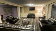This single-floor lounge space is designed for comfort and casual gatherings. The room centers around a generous seating arrangement featuring multiple dark leather sofas and armchairs, positioned around a round glass coffee table to create an inviting conversational setting. A wall-mounted flat-screen television serves as a focal point on the main wall, making the space well-suited for entertainment and group viewing. The floor is covered in a bold patterned carpet with a geometric design in muted tones of gold, black, and grey, adding a distinctive visual character to the room. The lower portion of the walls features white wainscoting, while the upper walls are painted in warm neutral tones, contributing to a cozy ambiance. A floor lamp in the corner provides soft supplemental lighting alongside the ceiling-mounted flush light fixture. A textured ceiling adds a classic touch overhead. The space also includes access to an adjacent area visible through an open doorway, suggesting additional utility or room. Horizontal blinds on the window allow for light control. Overall, this lounge offers a functional and well-appointed environment suitable for socializing, watching television, or unwinding in a relaxed setting.