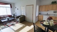 This single-story space is designed with an open-concept floor plan that seamlessly integrates the living, dining, and kitchen areas into one cohesive environment. The living area features light hardwood-style flooring and is furnished with comfortable seating arranged around a coffee table, anchored by a patterned area rug. A floor lamp and wall-mounted accent pieces add functional and decorative touches to the space. Natural light filters generously through several windows fitted with horizontal blinds, brightening the neutral-toned walls throughout.

The front entry is highlighted by a classic six-panel white door with wood trim, providing a clean and welcoming focal point. Adjacent to the entry, the kitchen area is outfitted with light wood cabinetry, light-colored countertops, and a dishwasher, offering practical storage and workspace. A small decorative arrangement on the counter adds a subtle homey detail.

The dining area, partially visible in the foreground, features a dark-toned table and chairs, providing a contrast to the lighter elements throughout the space. Overall, the layout feels open and functional, with a cohesive, understated aesthetic maintained across all areas.