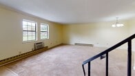 Located at 65 E Washington St in North Attleboro, MA, this two-floor residence offers a thoughtfully designed layout with a range of practical and attractive features throughout. The kitchen is well-equipped with white shaker-style cabinetry, dark granite countertops, a stone tile backsplash, warm hardwood flooring, and a full suite of stainless steel appliances including a refrigerator, range, dishwasher, and microwave. A carpeted living area on the lower level connects to the upper floor via a staircase with a black metal handrail, adding a clean architectural detail to the interior. The upper level opens to a spacious, light-filled common area with large windows offering views of the surrounding greenery, baseboard heating, and wall-unit climate control. The bedrooms are generously sized, carpeted, and feature double closets with paneled doors, providing ample storage. The bathrooms are finished with classic white subway tile wainscoting accented by decorative mosaic tile borders. The full bathroom includes a soaking tub, tile flooring, a wood vanity with a dark granite countertop, and a window allowing natural light. Soft yellow-toned walls and clean white trim carry consistently throughout, lending the space a cohesive, welcoming ambiance.