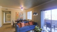 This single-floor residence offers a thoughtfully designed open-concept layout that seamlessly connects the living and dining areas into one cohesive, functional space. The living area is anchored by warm, neutral-toned walls accented with elegant crown molding that adds a refined architectural touch throughout. A ceiling fan with integrated lighting provides both comfort and ambiance overhead. Natural light filters in through a blinds-covered window, while a sliding glass door opens to the exterior, creating a bright and airy atmosphere. A striking arc floor lamp with a dome shade serves as a bold modern accent alongside the living area. The dining space is positioned adjacent to the living room, featuring a dark-toned dining table and chairs, complemented by decorative tableware and vibrant artwork on the wall — a colorful, abstract piece that adds personality and visual interest to the room. The overall aesthetic blends contemporary and eclectic elements, with warm earth tones, vivid accent colors, and layered lighting creating an inviting and comfortable ambiance throughout the space.