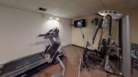 This single-floor fitness room is a practical, well-equipped exercise space designed to support a full range of workout activities. The room features concrete block walls finished in a neutral tan tone, a drop tile ceiling with recessed and track lighting, and durable low-pile carpeting throughout.

The exercise equipment is the centerpiece of the space. A Hoist multi-function cable and strength machine anchors one corner of the room, accompanied by a wall-mounted exercise chart for reference. A commercial-grade treadmill occupies another section of the room, offering cardiovascular training options, while a recumbent stationary bike provides a low-impact cycling alternative. Together, these machines offer a well-rounded selection of both strength and cardio equipment.

For comfort and entertainment during workouts, a flat-screen television is mounted on the wall alongside a wall-mounted oscillating fan to help circulate air. A framed fitness poster adds a motivational visual element to the space.

Access to the fitness room is provided through a standard door, with a view to an adjacent hallway or corridor visible from the interior. The overall ambiance is functional and utilitarian, with efficient use of the available footprint to maximize workout options in a compact, dedicated fitness environment.