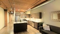 This single-floor loft space showcases a striking blend of industrial and contemporary design. The open-plan layout seamlessly connects the kitchen and living areas, creating a spacious and cohesive environment. Soaring ceilings with warm wood paneling and exposed metal ductwork define the industrial character of the space, while track lighting runs along the ceiling to provide ample, flexible illumination throughout.

The kitchen is a standout feature, outfitted with sleek dark cabinetry, light stone countertops, and a full suite of stainless steel appliances, including a refrigerator, range, microwave, and dishwasher. A central kitchen island with a dark base and matching countertop serves as both a functional prep area and a casual dining or gathering spot. A subway tile backsplash adds a clean, classic accent to the kitchen walls.

Light hardwood-style flooring flows throughout the open space, complementing the warm tones of the wood ceiling above. The living area transitions naturally from the kitchen, offering room for comfortable seating. Decorative greenery adds touches of natural life to the space. The overall ambiance is modern and urban, balancing raw industrial elements with refined finishes for a stylish and functional living environment.