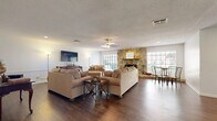 This well-appointed single-floor space in the United States offers a generous open-plan layout that seamlessly integrates the living and dining areas into one cohesive environment. The rich, dark hardwood flooring runs throughout, providing a warm and grounding foundation that contrasts beautifully with the bright white walls and textured ceiling.

The focal point of the room is a striking floor-to-ceiling natural stone fireplace, flanked by large windows that flood the space with natural light and offer views of the surrounding greenery. A wooden mantel tops the fireplace, adding a classic finishing touch. The ceiling fan centered overhead provides both comfort and visual balance to the room.

The living area is thoughtfully arranged with comfortable upholstered seating and an accent side table, creating an inviting atmosphere for relaxation. A wall-mounted flat-screen television is positioned opposite the seating, complemented by a traditional wooden console. A dining set with sleek metal chairs occupies the adjacent area near the windows, making it ideal for casual meals.

Recessed lighting and wainscoting details along the lower walls add architectural interest and a refined touch throughout. The open sightlines to additional areas of the property suggest further functional spaces beyond, enhancing the overall sense of depth and connectivity within this welcoming interior.