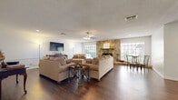 This well-appointed single-floor space in the United States offers a generous open-plan layout that seamlessly integrates the living and dining areas into one cohesive environment. The rich, dark hardwood flooring runs throughout, providing a warm and grounding foundation that contrasts beautifully with the bright white walls and textured ceiling.

The focal point of the room is a striking floor-to-ceiling natural stone fireplace, flanked by large windows that flood the space with natural light and offer views of the surrounding greenery. A wooden mantel tops the fireplace, adding a classic finishing touch. The ceiling fan centered overhead provides both comfort and visual balance to the room.

The living area is thoughtfully arranged with comfortable upholstered seating and an accent side table, creating an inviting atmosphere for relaxation. A wall-mounted flat-screen television is positioned opposite the seating, complemented by a traditional wooden console. A dining set with sleek metal chairs occupies the adjacent area near the windows, making it ideal for casual meals.

Recessed lighting and wainscoting details along the lower walls add architectural interest and a refined touch throughout. The open sightlines to additional areas of the property suggest further functional spaces beyond, enhancing the overall sense of depth and connectivity within this welcoming interior.