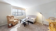 This single-floor unit offers a practical and functional layout across several distinct areas. The living room is carpeted and naturally lit by a wide window dressed with horizontal blinds, allowing ample daylight to fill the space. The room accommodates a seating area comfortably, with neutral walls contributing to a clean, understated aesthetic.

The kitchen is compact and efficiently arranged, featuring white cabinetry above and below the counter, a gas range, a refrigerator, and a single-basin sink. Light tile flooring and a microwave shelf add to the kitchen's utilitarian design.

There are two bedrooms, both carpeted and simply appointed. The larger bedroom benefits from elevated open shelving along the upper wall, a ceiling fan, and a closet with bifold doors, offering practical storage solutions. The second bedroom is more intimate in scale, with a window overlooking the exterior and a wall-mounted heating unit beneath it.

The bathroom features a classic retro aesthetic with pink ceramic wall tiles, a white vanity with a medicine cabinet mirror, and a shower-tub combination with a curtain. The overall ambiance of the unit is clean, neutral, and functional, suited for everyday living.