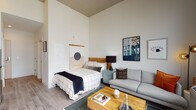 This thoughtfully designed studio unit offers an open-plan layout that efficiently combines the sleeping, living, and kitchen areas into a single cohesive space. Soaring ceilings amplify the sense of volume and airiness throughout, while light-toned hardwood-style flooring runs continuously underfoot, unifying the entire unit.

The living area features generous natural light streaming through large floor-to-ceiling windows and a glass door that opens onto a private balcony, offering open urban views of the surrounding cityscape. The sleeping area is defined by a wood-headboard bed frame and complementary wall art, creating a calm, curated atmosphere.

The kitchen is well-equipped with crisp white cabinetry extending to ceiling height, stainless steel appliances including a range, microwave, and refrigerator, a subway tile backsplash, and a built-in media console with integrated storage and display space for a television.

The bathroom is finished with neutral tile flooring and wall accents, featuring a glass-enclosed walk-in shower, a vanity with storage, and a clean, minimalist aesthetic consistent with the rest of the unit.

Throughout the space, the design balances functionality and modern style, making effective use of every square foot while maintaining a bright, welcoming ambiance.