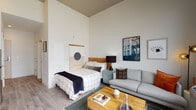 This thoughtfully designed studio unit offers an open-plan layout that efficiently combines the sleeping, living, and kitchen areas into a single cohesive space. Soaring ceilings amplify the sense of volume and airiness throughout, while light-toned hardwood-style flooring runs continuously underfoot, unifying the entire unit.

The living area features generous natural light streaming through large floor-to-ceiling windows and a glass door that opens onto a private balcony, offering open urban views of the surrounding cityscape. The sleeping area is defined by a wood-headboard bed frame and complementary wall art, creating a calm, curated atmosphere.

The kitchen is well-equipped with crisp white cabinetry extending to ceiling height, stainless steel appliances including a range, microwave, and refrigerator, a subway tile backsplash, and a built-in media console with integrated storage and display space for a television.

The bathroom is finished with neutral tile flooring and wall accents, featuring a glass-enclosed walk-in shower, a vanity with storage, and a clean, minimalist aesthetic consistent with the rest of the unit.

Throughout the space, the design balances functionality and modern style, making effective use of every square foot while maintaining a bright, welcoming ambiance.