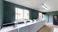 This single-floor laundry facility is designed with practicality and efficiency in mind. The space features a linear arrangement of commercial-grade, coin-operated washing machines and dryers lined along one wall, providing ample capacity for multiple users simultaneously. The machines are uniformly white, lending a clean and organized aesthetic to the room.

The walls are painted in a deep teal-green tone, creating a distinct and modern visual contrast against the white appliances and bright ceiling. Overhead, fluorescent lighting ensures the space is well-lit throughout. Large sliding windows along the main wall allow natural light to filter in, offering views of a landscaped exterior with neatly trimmed hedges, further contributing to the bright and airy atmosphere.

The flooring is a neutral, light-toned surface that complements the overall clean and utilitarian design. A utility sink and additional equipment are visible at one end of the machine row, adding functional versatility to the space. The layout is open and uncluttered, allowing for easy movement and access to all appliances. Overall, the facility presents a tidy, well-maintained environment suited for convenient and efficient everyday use.