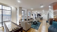 This contemporary single-floor space showcases a thoughtfully designed open-concept layout that seamlessly connects its key living areas. The main living zone features a neutral-toned seating arrangement centered around a bold teal area rug, complemented by a dark wood coffee table and tasteful decorative accents. Structural columns add architectural character while defining the flow between spaces. Large, multi-pane windows along the perimeter flood the interior with abundant natural light and offer views of the surrounding greenery, enhancing the bright and airy ambiance throughout. Warm hardwood flooring runs continuously across the space, adding a sense of cohesion and elegance. A shuffleboard table positioned near the entrance introduces a recreational element, making the space as functional as it is inviting. Toward the rear, a well-appointed kitchen features dark cabinetry, a tile backsplash, stainless steel appliances, and a kitchen island with bar seating illuminated by pendant lighting. Recessed lighting throughout ensures a well-lit environment at all times. The overall atmosphere is modern and sophisticated, striking a balance between comfort, style, and practicality.