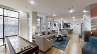 This contemporary single-floor space showcases a thoughtfully designed open-concept layout that seamlessly connects its key living areas. The main living zone features a neutral-toned seating arrangement centered around a bold teal area rug, complemented by a dark wood coffee table and tasteful decorative accents. Structural columns add architectural character while defining the flow between spaces. Large, multi-pane windows along the perimeter flood the interior with abundant natural light and offer views of the surrounding greenery, enhancing the bright and airy ambiance throughout. Warm hardwood flooring runs continuously across the space, adding a sense of cohesion and elegance. A shuffleboard table positioned near the entrance introduces a recreational element, making the space as functional as it is inviting. Toward the rear, a well-appointed kitchen features dark cabinetry, a tile backsplash, stainless steel appliances, and a kitchen island with bar seating illuminated by pendant lighting. Recessed lighting throughout ensures a well-lit environment at all times. The overall atmosphere is modern and sophisticated, striking a balance between comfort, style, and practicality.