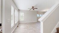 This two-story home offers a well-organized layout spread across two floors, with the main level housing the living, dining, and kitchen areas, and the upper level dedicated to the bedrooms and additional bathrooms. The main level greets you with a bright, open living area finished in light wood-style flooring and neutral warm-gray walls. Large windows with blinds invite abundant natural light, while a ceiling fan overhead adds both style and function. A staircase with white balustrade connects the two floors, and a row of elevated windows frames scenic views of surrounding hills. The kitchen features dark cabinetry with dark countertops, light wood-style flooring, recessed lighting, and stainless steel appliances including a full-size refrigerator and dishwasher. A half bathroom on the main level includes a classic pedestal sink with chrome fixtures and a mirror. Upstairs, the generously sized primary bedroom is carpeted and includes a walk-in closet with built-in shelving and a ceiling fan with multiple windows allowing natural light. The second bedroom is also carpeted, with a ceiling fan and a multi-panel window arrangement. Three bathrooms in total serve the household comfortably across both floors.