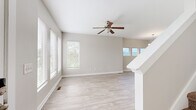 This two-story home offers a well-organized layout spread across two floors, with the main level housing the living, dining, and kitchen areas, and the upper level dedicated to the bedrooms and additional bathrooms. The main level greets you with a bright, open living area finished in light wood-style flooring and neutral warm-gray walls. Large windows with blinds invite abundant natural light, while a ceiling fan overhead adds both style and function. A staircase with white balustrade connects the two floors, and a row of elevated windows frames scenic views of surrounding hills. The kitchen features dark cabinetry with dark countertops, light wood-style flooring, recessed lighting, and stainless steel appliances including a full-size refrigerator and dishwasher. A half bathroom on the main level includes a classic pedestal sink with chrome fixtures and a mirror. Upstairs, the generously sized primary bedroom is carpeted and includes a walk-in closet with built-in shelving and a ceiling fan with multiple windows allowing natural light. The second bedroom is also carpeted, with a ceiling fan and a multi-panel window arrangement. Three bathrooms in total serve the household comfortably across both floors.