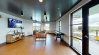 Located at 2880–2888 NE Holladay St in Portland, Oregon, this contemporary community game room is thoughtfully designed for recreation and social engagement. The space features warm wood-look tile flooring throughout and a striking dark ceiling that contrasts with the light-toned walls, creating a modern, energetic atmosphere.

The main gaming area is generously proportioned and anchored by a foosball table and a full-length shuffleboard table, offering ample room for gameplay and spectators alike. A dedicated alcove with bold orange accent walls provides an immersive nook housing a classic arcade cabinet and a dartboard, adding a retro-inspired touch to the overall design. Teal accent trim borders the transition between zones, giving the space a cohesive and lively color palette.

A comfortable lounge area with mid-century modern seating and a wall-mounted flat-screen TV invites relaxation between games. A storage console with organized bins adds functional charm. Pendant and recessed lighting throughout ensures the space is well-lit at any hour.

Multiple large windows flood the interior with natural light and frame views of the surrounding Portland skyline and greenery. French doors open directly onto an outdoor terrace, extending the social space outside. The overall ambiance is dynamic yet welcoming, making it an ideal destination for community gathering and casual entertainment.