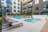 Located at 401 N Main St in the heart of Santa Ana, California, this outdoor amenity courtyard is a standout feature of a modern multi-story residential community. The space is defined by its resort-style swimming pool, which serves as the focal point of the courtyard and is surrounded by a generous tiled sun deck outfitted with contemporary lounge chairs and wooden daybeds, ideal for relaxing in the Southern California sunshine.

The courtyard is enclosed by the building's multi-story façade, which blends contrasting grey and white exterior panels with large windows and private balconies that overlook the amenity level below. Lush tropical landscaping, including tall palm trees, banana leaf plants, agave, and an array of potted succulents and ornamental grasses, lends a vibrant, resort-like atmosphere to the space.

A pergola-covered lounge area with pendant globe lighting provides a shaded retreat alongside the pool, while a separate green lawn area with a wooden ping-pong table and curated planting beds offers additional recreational space. Floor-to-ceiling glass walls on the ground level connect interior amenity spaces visually to the outdoor courtyard. The overall design balances modern urban architecture with warm, inviting outdoor living elements, creating a cohesive and well-appointed communal environment.