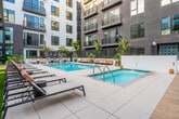 Located at 401 N Main St in the heart of Santa Ana, California, this outdoor amenity courtyard is a standout feature of a modern multi-story residential community. The space is defined by its resort-style swimming pool, which serves as the focal point of the courtyard and is surrounded by a generous tiled sun deck outfitted with contemporary lounge chairs and wooden daybeds, ideal for relaxing in the Southern California sunshine.

The courtyard is enclosed by the building's multi-story façade, which blends contrasting grey and white exterior panels with large windows and private balconies that overlook the amenity level below. Lush tropical landscaping, including tall palm trees, banana leaf plants, agave, and an array of potted succulents and ornamental grasses, lends a vibrant, resort-like atmosphere to the space.

A pergola-covered lounge area with pendant globe lighting provides a shaded retreat alongside the pool, while a separate green lawn area with a wooden ping-pong table and curated planting beds offers additional recreational space. Floor-to-ceiling glass walls on the ground level connect interior amenity spaces visually to the outdoor courtyard. The overall design balances modern urban architecture with warm, inviting outdoor living elements, creating a cohesive and well-appointed communal environment.