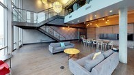 This sophisticated urban space offers an exceptional blend of industrial design and modern elegance, set high above the St. Louis skyline with breathtaking views of the Gateway Arch and surrounding downtown cityscape. Floor-to-ceiling glass curtain walls span the perimeter, flooding the interior with natural light and framing the iconic cityscape as a living backdrop.

The main living and lounge area features soaring double-height ceilings anchored by dramatic architectural columns and an eye-catching industrial steel staircase with glass railings leading to an open mezzanine level above. Sculptural artichoke-style pendant chandeliers hang at varying heights, adding warmth and artistic flair to the expansive open floor plan. Wide-plank hardwood flooring runs throughout, complementing the raw, textured wall treatments.

Flexible seating arrangements include generous contemporary sofas and accent chairs positioned around a round coffee table, ideal for social gatherings. A long communal dining table lined with modern white chairs provides dedicated space for group meals or meetings. An exposed concrete ceiling adds to the industrial character of the space.

A separate lounge area near the glass facade offers intimate seating with direct views of the Arch, city buildings, and outdoor terrace, making it a standout destination for events or private gatherings in the heart of St. Louis.