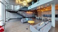 This sophisticated urban space offers an exceptional blend of industrial design and modern elegance, set high above the St. Louis skyline with breathtaking views of the Gateway Arch and surrounding downtown cityscape. Floor-to-ceiling glass curtain walls span the perimeter, flooding the interior with natural light and framing the iconic cityscape as a living backdrop.

The main living and lounge area features soaring double-height ceilings anchored by dramatic architectural columns and an eye-catching industrial steel staircase with glass railings leading to an open mezzanine level above. Sculptural artichoke-style pendant chandeliers hang at varying heights, adding warmth and artistic flair to the expansive open floor plan. Wide-plank hardwood flooring runs throughout, complementing the raw, textured wall treatments.

Flexible seating arrangements include generous contemporary sofas and accent chairs positioned around a round coffee table, ideal for social gatherings. A long communal dining table lined with modern white chairs provides dedicated space for group meals or meetings. An exposed concrete ceiling adds to the industrial character of the space.

A separate lounge area near the glass facade offers intimate seating with direct views of the Arch, city buildings, and outdoor terrace, making it a standout destination for events or private gatherings in the heart of St. Louis.