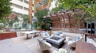 This thoughtfully designed outdoor courtyard amenity space offers a compelling blend of modern architecture and resort-style living. The centerpiece of the main deck is a sleek, square fire pit surrounded by contemporary wicker lounge chairs and accent pillows in crisp white and blue, creating an inviting gathering area. A ping pong table adds a recreational element to the deck, which is finished in warm horizontal hardwood planking. A striking curved wooden slatted pergola structure provides architectural interest and partial shade, while abundant planters and mature trees throughout the courtyard enhance the sense of a lush, verdant retreat. An adjacent spa pool, tiled in cool blue mosaic, is flanked by lounge chairs and framed by tropical plants, offering a tranquil space to unwind. Steps away, a built-in hot tub features a sculptural Corten steel waterfall element set against a concrete tile wall, lending an industrial-chic aesthetic. Concrete stairways and pathways connect the various levels of the courtyard, leading to building interiors framed by steel-accented windows. Multi-story balconies with warm wood railings overlook the courtyard, tying the outdoor amenity space cohesively to the surrounding building's contemporary design language.