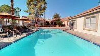 This inviting outdoor amenity area showcases a generously sized swimming pool with crystal-clear turquoise water, perfect for relaxation and recreation. The pool is complemented by a separate circular spa, ideal for unwinding, and the entire area is finished with attractive paver stonework that extends across the expansive sun deck. Lounge chairs and shaded umbrella seating are arranged throughout the deck, offering plenty of options for both sun-soaking and sheltered relaxation. A dedicated pool house structure, finished in a stucco exterior with a red tile roof in a classic Southwestern architectural style, provides convenient amenities including a vending area. The surrounding landscape is lush with towering palm trees and mature shade trees that frame the space beautifully, creating a private, resort-like atmosphere. The overall ambiance is bright, open, and sun-drenched, consistent with a warm desert climate, making this outdoor space a standout communal feature well-suited for year-round enjoyment.