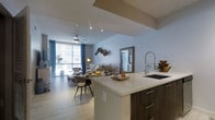This single-floor space showcases a thoughtfully designed open-concept layout that seamlessly connects the kitchen, dining, and living areas into one cohesive environment. The kitchen features a large central island with a crisp white countertop and dark wood cabinetry, complemented by a professional-style pull-down faucet and an undermount sink. Recessed lighting illuminates the space from a tray ceiling detail overhead, adding architectural interest.

The living area extends naturally from the kitchen, anchored by a plush neutral-toned sofa arrangement and accented by bold teal drapery panels that frame a generous window allowing abundant natural light to fill the room. A ceiling fan overhead adds both function and style. Light wood-tone flooring runs throughout, contributing to the warm, airy ambiance. A decorative mirror and wall art enhance the living space, while a small dining area with modern chairs bridges the kitchen and lounge zones. The overall design balances contemporary finishes with comfortable, livable details.