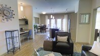 This single-floor residence showcases an open-concept design that integrates the living, dining, and kitchen areas into one cohesive, flowing space. Hardwood-style flooring runs throughout, adding warmth and visual continuity across all zones.

The living area is anchored by a dark leather armchair paired with a woven ottoman, creating a comfortable and stylish seating arrangement. A glass-topped coffee table adds a contemporary touch, while a neutral area rug defines the space.

The kitchen features a dark-toned island with a built-in sink, stainless steel appliances, and pendant lighting overhead. French doors with surrounding windows invite natural light into the kitchen and dining area, enhancing the open, airy feel of the space.

The dining area accommodates a full table with seating for multiple guests, positioned on a area rug beneath a flush-mount light fixture. A built-in desk or workspace is tucked along one wall, offering a functional nook for productivity.

Neutral gray walls, white curtains, and track lighting contribute to a clean, modern aesthetic throughout. The layout maximizes both functionality and comfort, making efficient use of the available space.