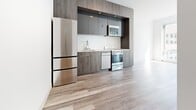 Located at 1177 Market St in the heart of San Francisco, this modern single-floor unit offers a clean, contemporary design throughout. Light wood-tone flooring runs seamlessly across all areas, lending the space a warm yet minimalist aesthetic.

The kitchen is a standout feature, outfitted with a full suite of stainless steel appliances including a refrigerator, range, dishwasher, and built-in microwave. Sleek two-tone cabinetry in gray and wood-grain finishes extends to the ceiling, maximizing storage while contributing to the sophisticated, modern look. A white tile backsplash and undermount sink complete the well-appointed cooking area.

The living and sleeping areas are bright and airy, bathed in natural light streaming through oversized floor-to-ceiling windows. These large windows frame urban views of the surrounding San Francisco cityscape, connecting the interior to the vibrant neighborhood outside. White walls throughout amplify the sense of openness and provide a neutral canvas.

A hallway connects the main living areas to additional rooms, maintaining a logical and efficient layout. The overall ambiance of the space is clean, modern, and light-filled, making it well-suited for those seeking a contemporary urban lifestyle in one of San Francisco's most central and accessible neighborhoods.